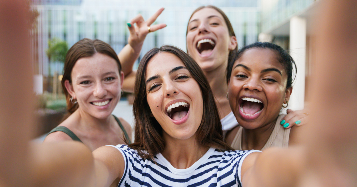 Invisalign - Four joyful women take a selfie outdoors, smiling widely and showing peace signs, creating a cheerful and vibrant atmosphere.