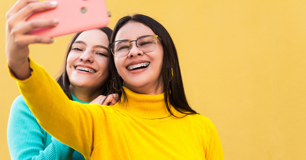 Two young women with braces take a cheerful selfie. One wears a yellow sweater, the other blue, against a vibrant yellow background. Their expressions are joyful.