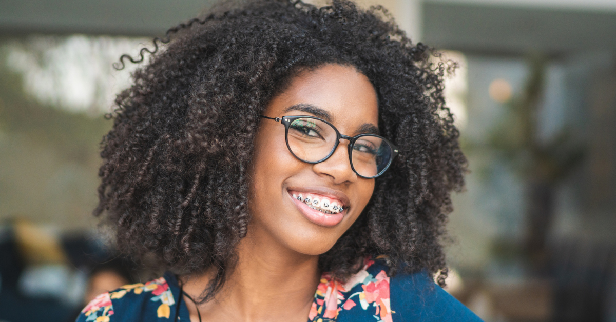 Smiling person with curly hair and glasses, wearing braces, in a floral top. The background is softly blurred, conveying a warm and cheerful tone.